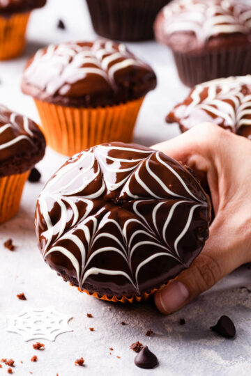 a hand holding a single chocolate fudge spiderweb muffin with some more muffins in the background.