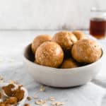 A straight-on angle shot of small bowl filled with coconut and peanut butter spiced energy bites with small jar of maple syrup in the background