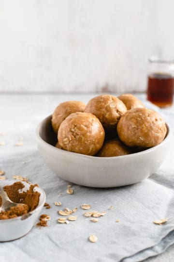 A straight-on angle shot of small bowl filled with coconut and peanut butter spiced energy bites with small jar of maple syrup in the background