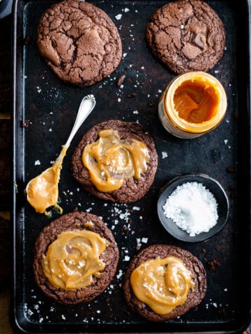 Overhead shot of five big chocolate salted caramel cookies on a baking tray with small bowl of sea salt and small jar of caramel