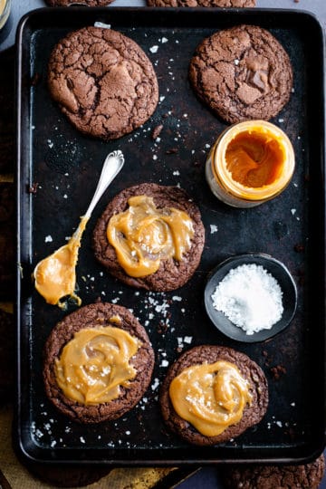 Overhead shot of five big chocolate salted caramel cookies on a baking tray with small bowl of sea salt and small jar of caramel