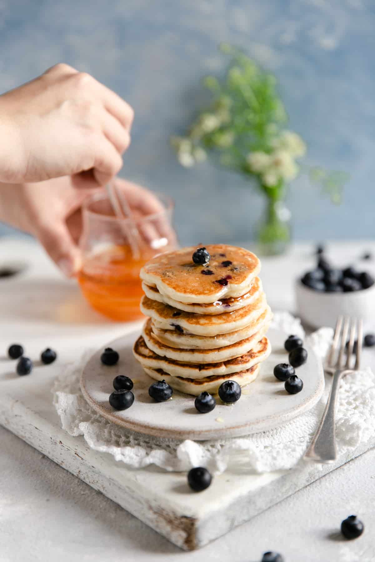 Straight ahead shot of small stack of blueberry pancakes with someone holding glass jar with agave nectar in the background