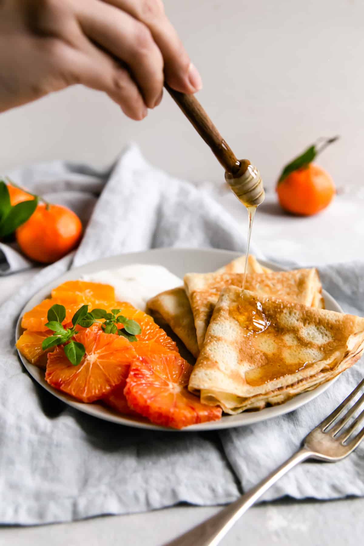 a person drizzling some homey over a plate with crepes and citrus