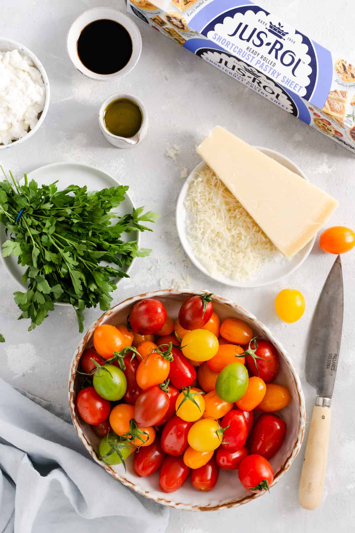 Overhead shot of the ingredients for mixed tomato and cheese tartlets: pastry, mixed baby plum tomatoes, ricotta, hard cheese, parsley, olive oil and balsamic glaze