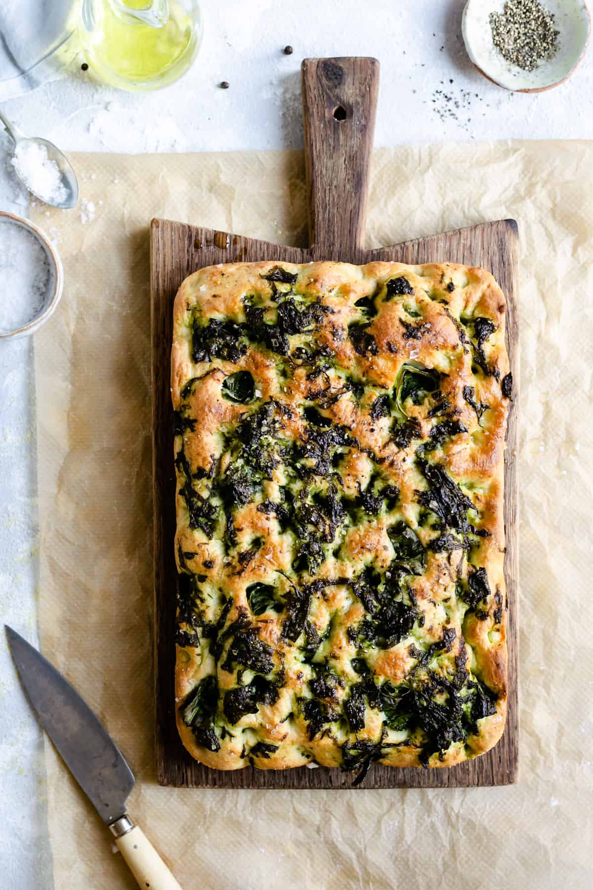 Overhead shot of a whole baked wild garlic and black pepper focaccia on a wooden chopping board with small knife on side