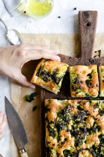 overhead shot of a person's hand holding a slice of wild garlic and black pepper focaccia with the rest of focaccia resting on a wooden chopping board