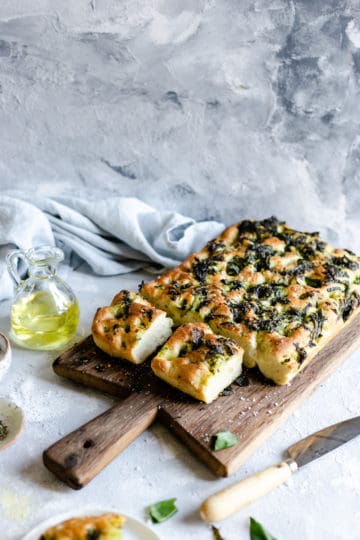 side angle of wild garlic focaccia on a wooden chopping board with few slices being cut out