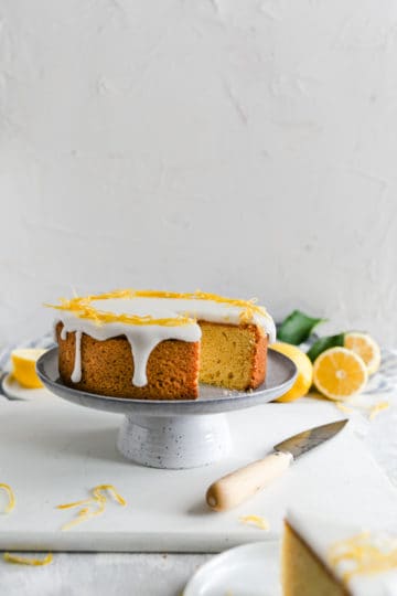 a straight ahead angle shot of a zesty lemon cake on a cake stand with one piece cut out