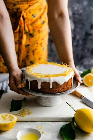 a side angle of a person holding a plate with whole lemon cake topped with candied lemon peel