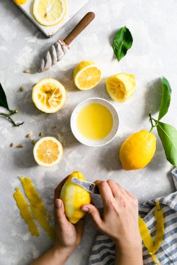 overhead shot of a person peeling a lemon with a small bowl of lemon juice and lemon halves on side