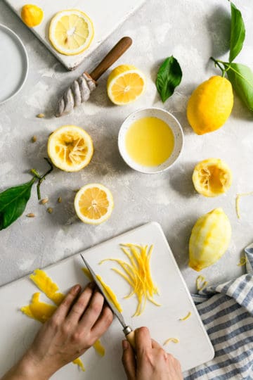 overhead shot of a person slicing lemon peel on a white chopping board surrounded with lemon halves