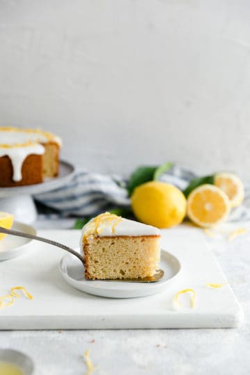 a side angle shot of one slice of zesty lemon cake on a small plate with fresh lemons in soft focus at the back