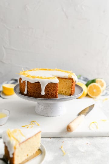 a straight ahead angle shot of a zesty lemon cake on a cake stand with one piece cut out