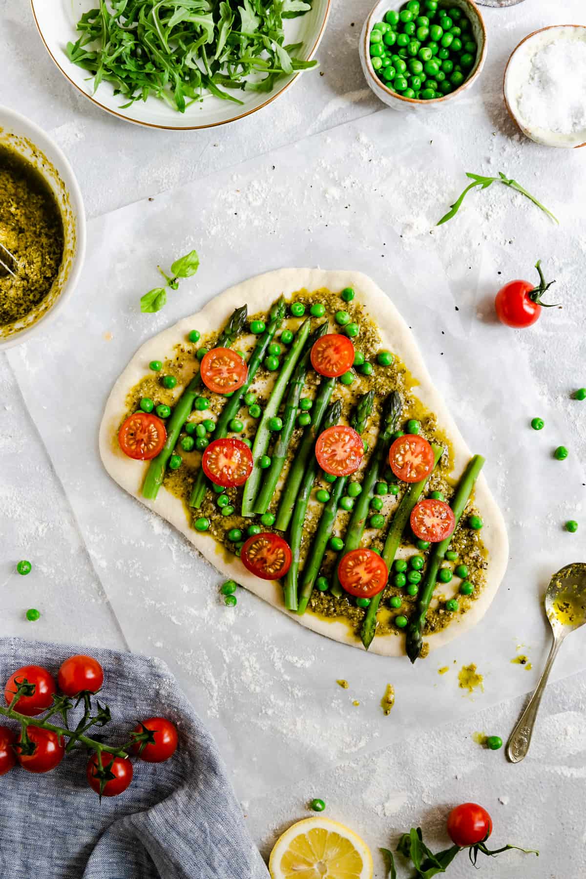 Top view of pizza dough topped with green pesto, asparagus, green peas and cherry tomatoes