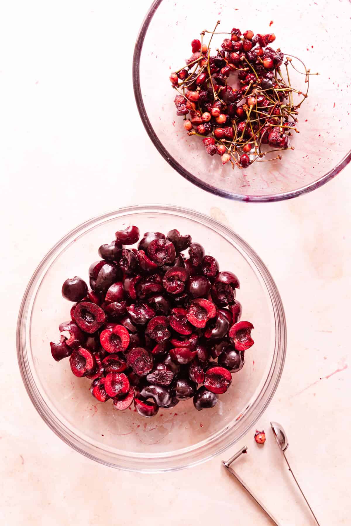 top view of pitted cherries in a bowl with another bowl of cherry stones and stalks