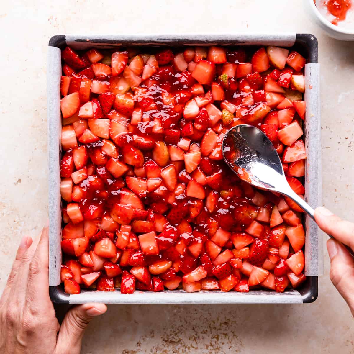 baking tin with chopped strawberries being spread on top of crumble base with spoon.