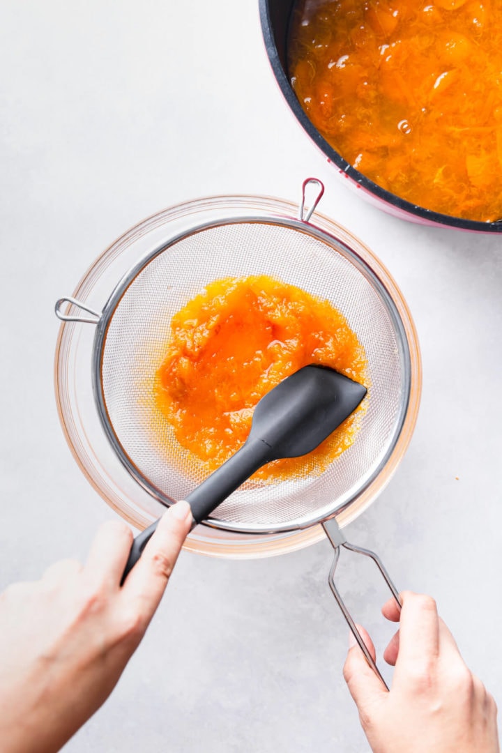 overhead view of a person pushing cooked apricots through the fine metal sieve into a bowl