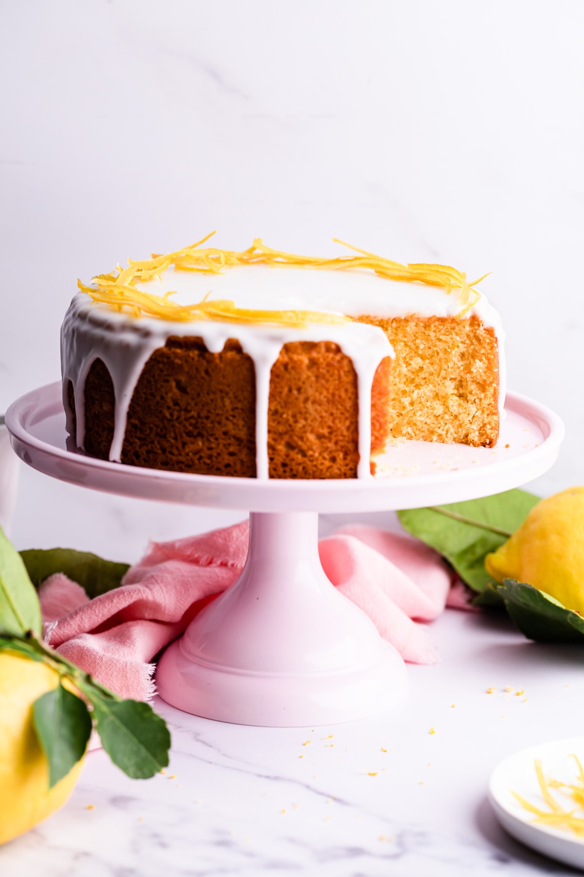 lemon cake with slice cut out on a pink cake stand and pink napkin behind it.
