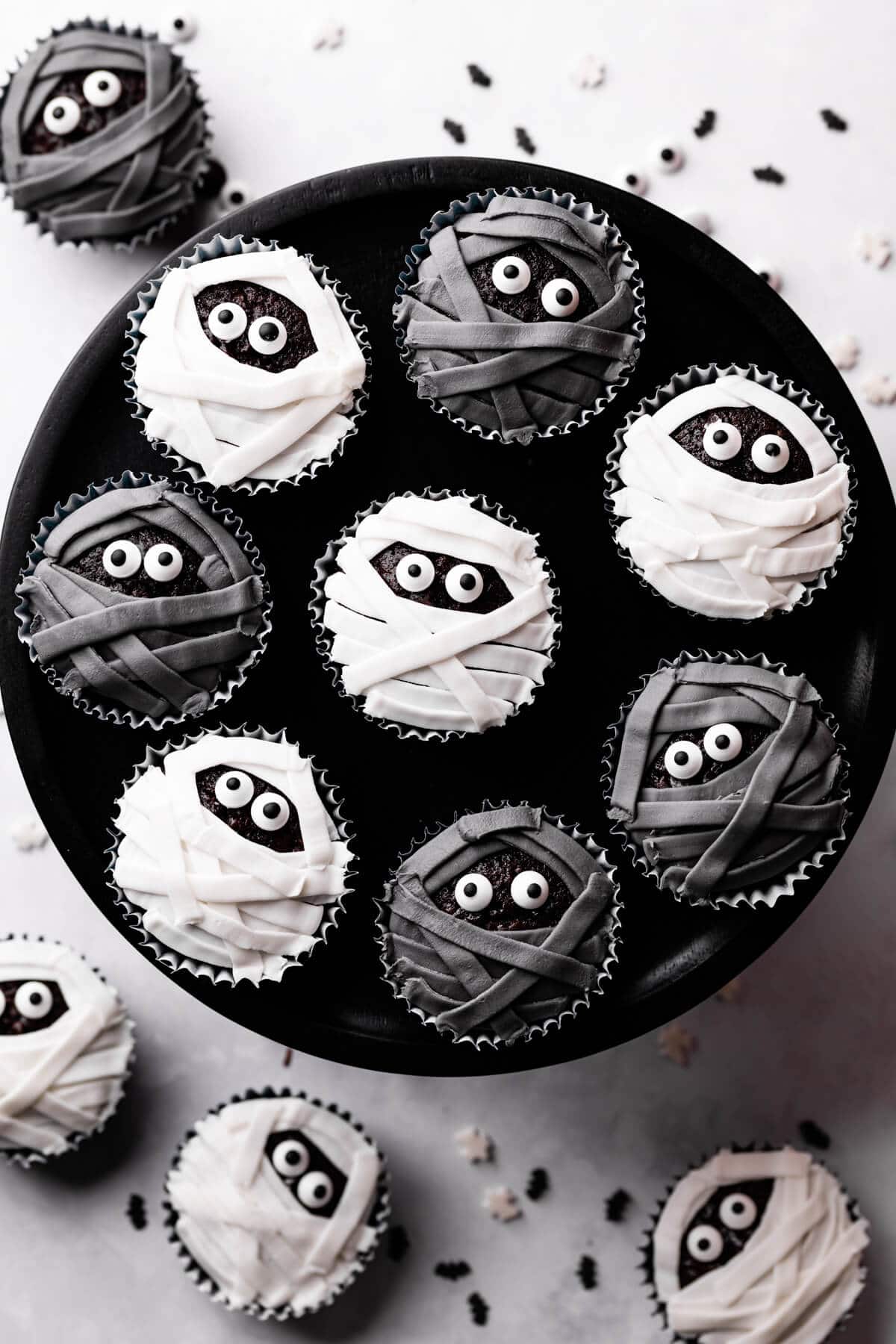 halloween mummy cupcakes on a black cake stand and sprinkles around it.