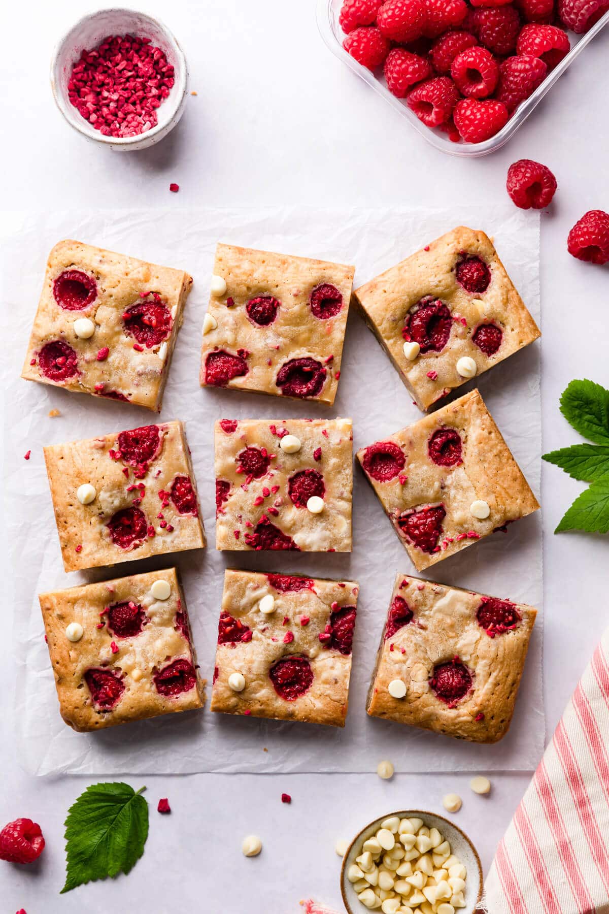 9 square slices of blondie bars with raspberries and white chocolate on a white surface.