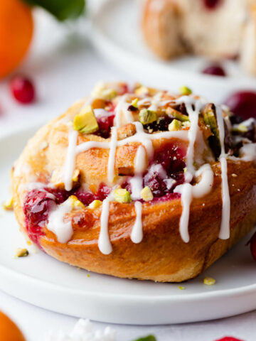 an individual orange and cranberry Chelsea bun on a small plate drizzled with icing.