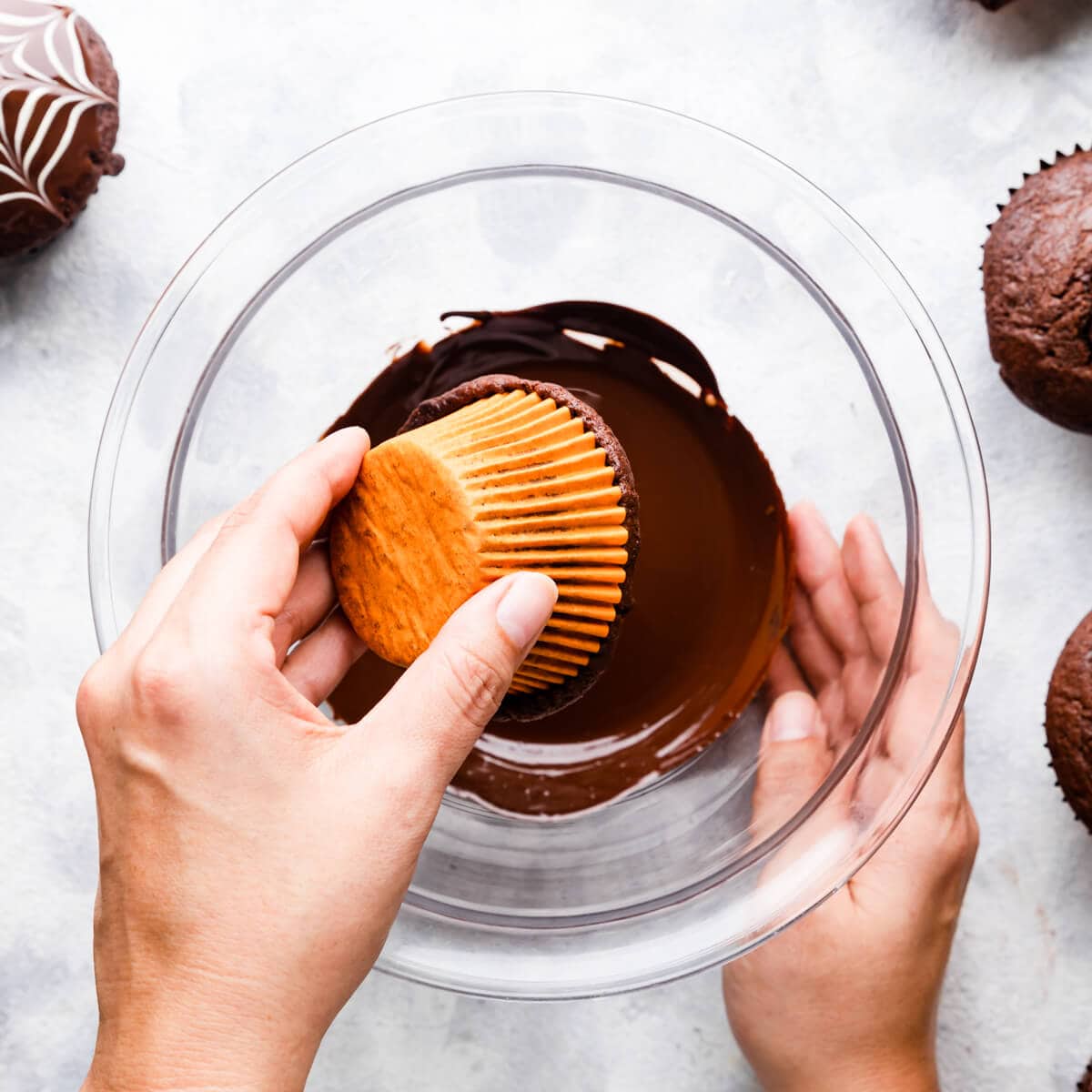 pair of hands dipping a chocolate muffin into a bowl with melted chocolate.