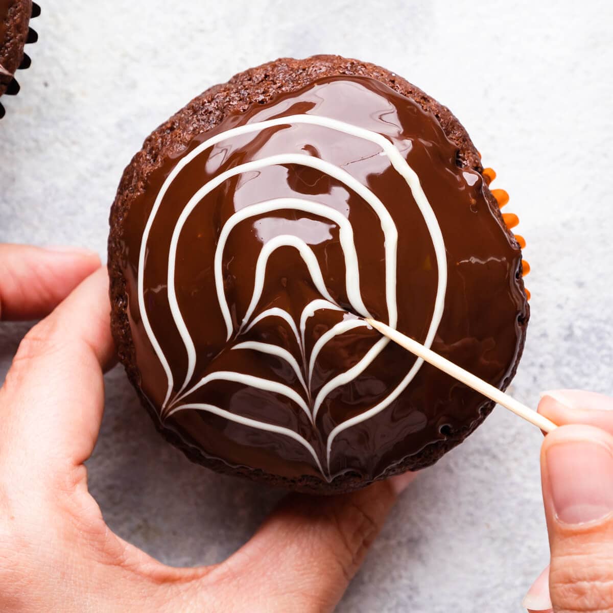 chocolate muffin being decorated with toothpick to create spiderweb design.