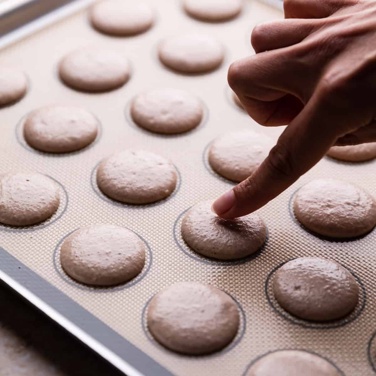 index finger touching gingerbread macaron piped on silicone baking mat.