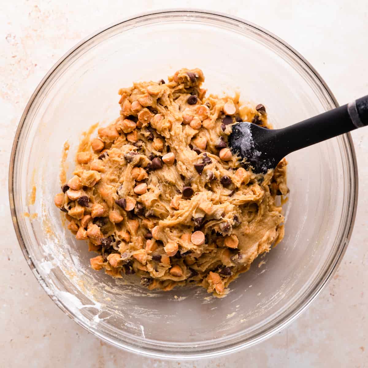 glass mixing bowl with cookie batter with butterscotch and chocolate chips inside and a rubber spatula inside of the bowl.