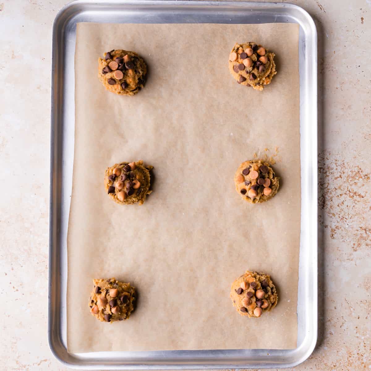 large metal baking sheet lined with brown paper and 6 portions of butterscotch cookies dough on it.
